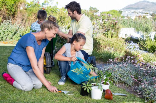 A family of four gardening in a well-maintained backyard, with two adults and two children, engaged in planting and tending to the garden. The foreground features a lush, green lawn with a young girl using a small trowel to plant seedlings in the soil, while her mother, dressed in a blue top and grey trousers, assists with the planting process. Nearby, a young boy in a striped shirt and his father, who is wearing a light-colored shirt, stand together, observing or participating in the activity. The garden includes flower beds edged with various flowering plants and shrubs, adding natural colour and texture to the scene. Gardening tools, small pots, and watering cans are visible around the area, indicating ongoing outdoor maintenance. The background reveals a scenic view of distant town rooftops and rolling hills, suggesting a suburban location consistent with herne hill or nearby areas. The weather is bright and clear, with natural sunlight highlighting the vibrant plant life and the relaxed, outdoor family activity, aligning with professional gardening and landscaping services aimed at creating inviting outdoor spaces in the UK.