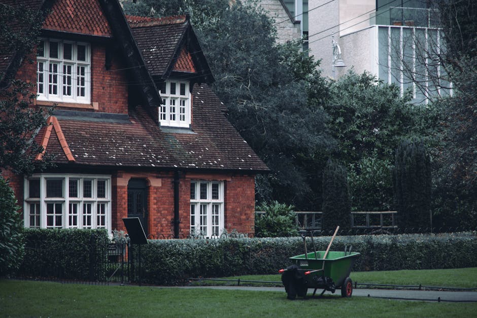 A small front garden in front of a traditional red-brick house with a steep, tiled roof and white-framed windows. The garden features a well-maintained lawn bordered by a low hedge, with a seating are