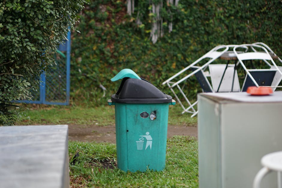 A green outdoor waste bin with a black lid and a light blue lid extension, positioned on a patch of grass near a garden with dense green bushes and trees in the background. The bin features a white gr