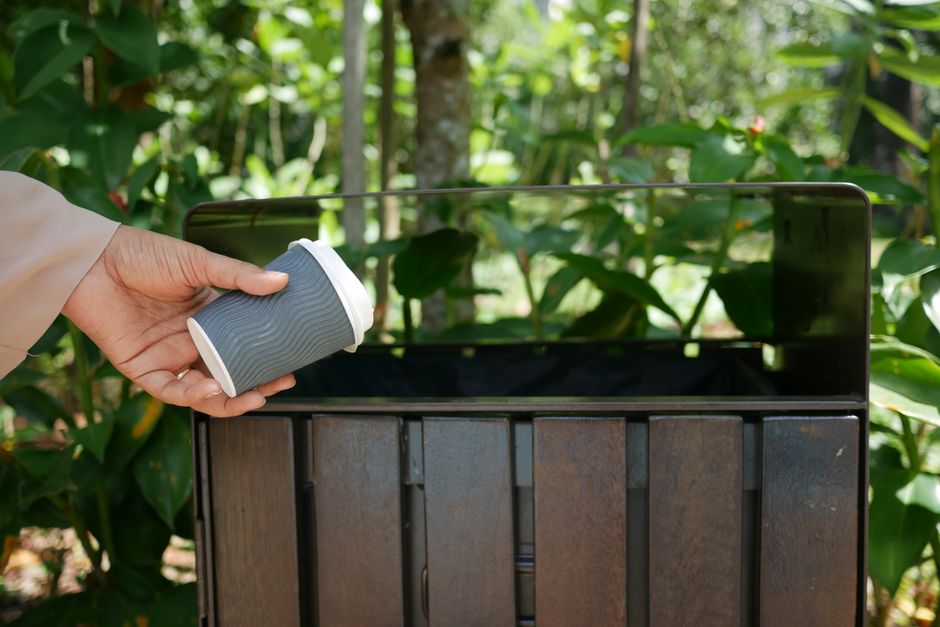 A green outdoor waste bin with a black lid and a light blue lid extension, positioned on a patch of grass near a garden with dense green bushes and trees in the background. The bin features a white gr